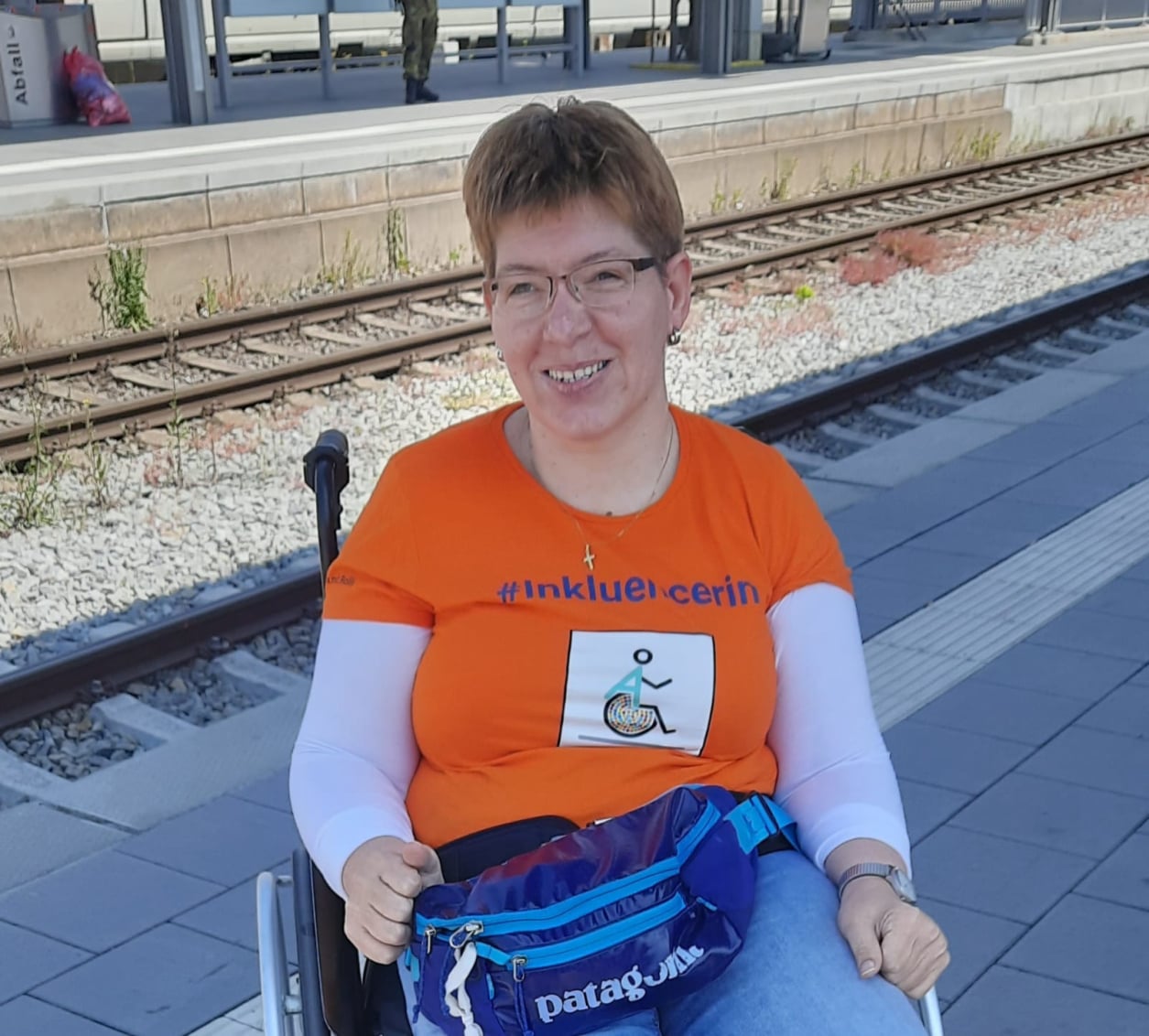 Evi sitzt im Rollstuhl am Bahnsteig und trägt ein T-Shirt mit Aufschrift "Inkluencerin".
